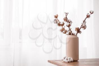 Vase with cotton branches on table near window�