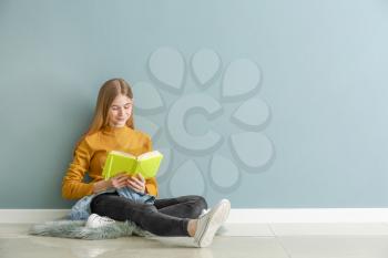 Cute teenage girl with book sitting on floor near color wall�
