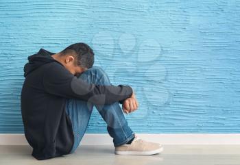 Sad African-American teenage boy sitting on floor near color wall�