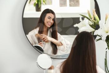 Beautiful young woman combing her healthy long hair near mirror at home�