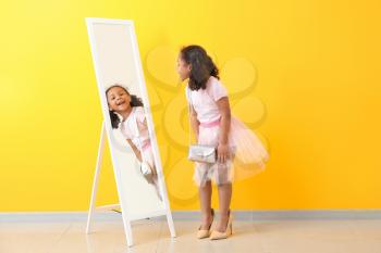 Adorable little African-American girl with mother's shoes looking into mirror near color wall�