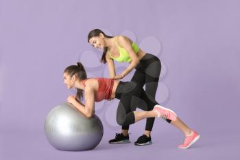 Young sporty woman with fitball doing exercises under supervision of her personal trainer on color background�