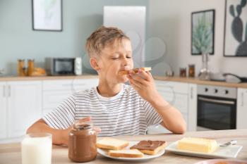 Funny little boy eating tasty toasts with chocolate spreading in kitchen�
