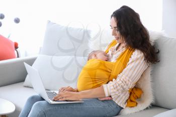 Young mother with little baby in sling using laptop at home�