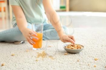 Careless little girl eating nuts and drinking juice while sitting on carpet�