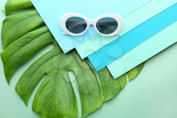 Stylish sunglasses with papers and tropical leaf on color background�