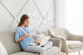 Beautiful pregnant woman with laptop sitting on sofa at home�