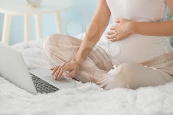 Beautiful pregnant woman with laptop sitting on bed�