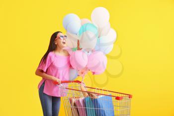 Young woman with shopping cart and balloons on color background�