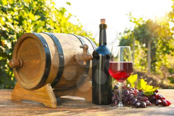 Glass and bottle of red wine with fresh grapes and barrel on wooden table in vineyard�