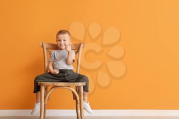 Portrait of cute little boy sitting on chair and showing thumb-up gesture against color background�
