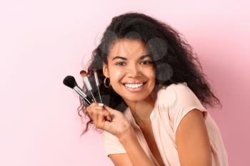 Beautiful African-American woman with makeup brushes on color background�