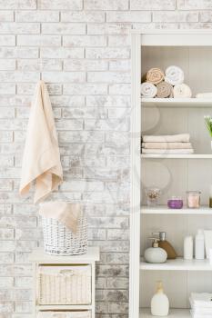 Shelf unit with towels and cosmetics near brick wall in bathroom�