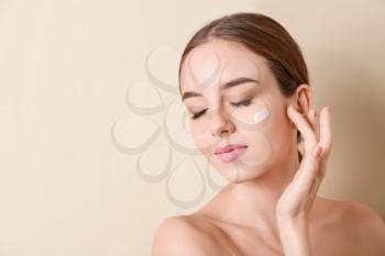 Beautiful young woman applying cream on her face against color background�