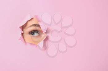 Eye of beautiful young woman visible through hole in pink teared paper�