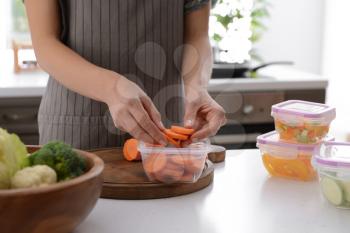 Woman putting fresh carrot slices into plastic container for freezing at table in kitchen�