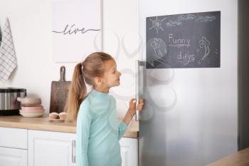 Little girl near chalkboard on refrigerator in kitchen�