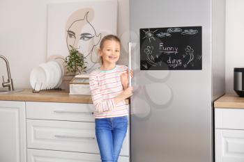 Little girl near chalkboard on refrigerator in kitchen�