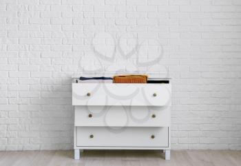 Modern chest of drawers with clothes near brick wall in room�