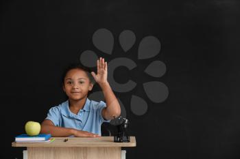 Little African-American pupil sitting at school desk against dark background�