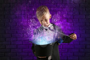 Cute little magician showing trick with hat against dark brick wall�