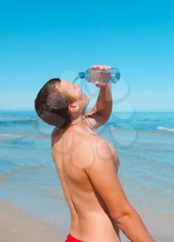muscular young man on the beach pouring down cold water on his face from plastic bottle