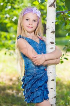 Nice little girl in jeans dress posing near birch