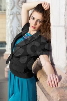 Curly brunette wearing blue dress and black jacket posing outdoors