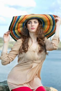 Pretty curly brunette in colorful hat sitting on a rock by the river