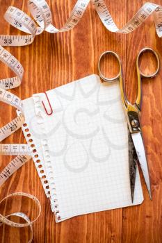 Scissors and tape measure on wooden background with empty paper sheet