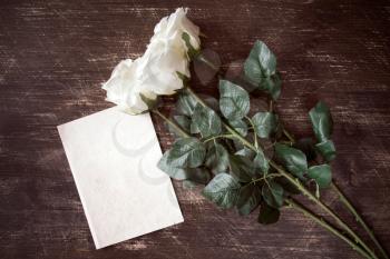 Blank paper card with artificial white roses on wooden background