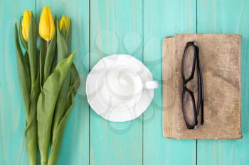 Old book with glasses,yellow tulips and empty cup on wooden background