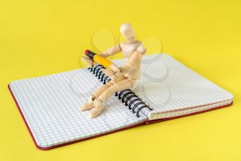 Wooden mannequin with a pencil sitting on a notebook.