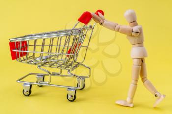 Wooden man pushing a shopping cart on the yellow background. Go for shopping concept.