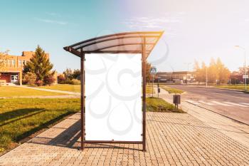 Vertical blank white billboard at bus stop on city street. In the background buildings and road. Mock up. Poster on street next to roadway. Sunny summer day.