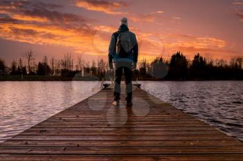 Silhouette of man standing on the lake wooden pier at sunset, human strength, psychology concept.Back view of adult man standing on bridge.