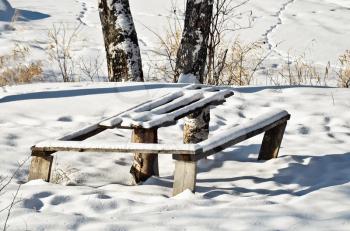 Table and bench, covered with white snow against a background of snow-covered meadows, trees, dry grass, trails