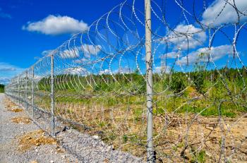 Barbed wire fence on a background of blue sky with white clouds, green trees, yellow soil and gravel