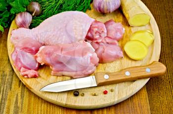 Cut pieces of chicken legs on a round board, garlic, parsley, ginger, dill, pepper and a knife on a wooden boards background