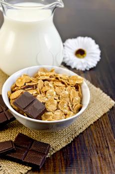 Corn flakes with slices of chocolate, milk in glass jug, hessian cloth, white flower on a wooden board