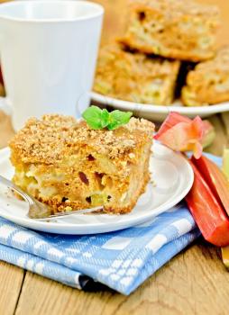 A piece of sweet cake with rhubarb, mint, blue napkin, cup on the background of wooden boards