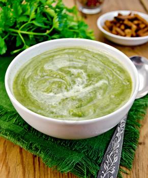 Green soup puree in a white bowl with spoon on a napkin, parsley, croutons, peppers on a wooden boards background