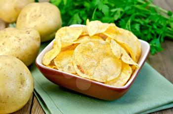 Potato chips in a clay bowl on a napkin, fresh potatoes, parsley on a wooden boards background