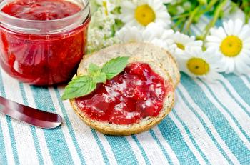Two slices of bread with strawberry jam, a jar of jam, a knife and a bouquet of daisies on a green striped napkin