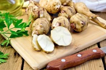 One cut and a few whole tubers of Jerusalem artichoke with parsley, knife and vegetable oil decanter on a wooden board
