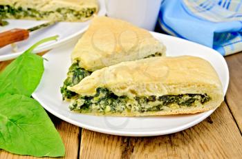 Two pieces of pie with spinach and cheese on a plate, spinach leaves, cup, knife, napkin on wooden board
