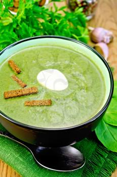 Green soup puree in bowl with a spoon on napkin, parsley, spinach, croutons, garlic and pepper on wooden board