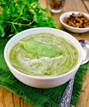 Green soup puree in white bowl with spoon, napkin, parsley, croutons, peppers on a wooden boards background