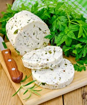 Homemade cheese with herbs and spices, cut into slices, knife, parsley, rosemary, basil, napkin on wooden board