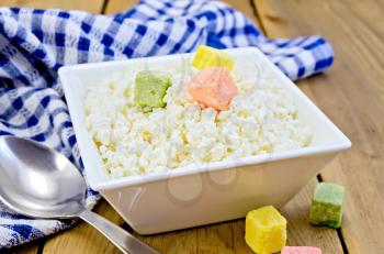 Cottage cheese in a white square bowl with colored sugar cubes, blue checkered napkin, spoon on a wooden board